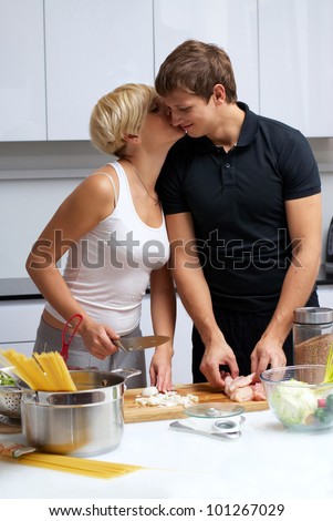 Playful young couple in their kitchen making dinner. kitchen is white and filled with light. couple is young and fit. she slices onion and kisses boyfriend