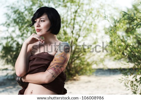 beautiful black haired girl with tattoo wrapped in brown cloth stands in the tree shade in a desert