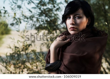 beautiful black haired girl wrapped in brown cloth stands in the tree shade in a desert