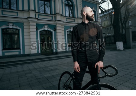 bearded caucasian bald man stands with black fix bicycle near old restored building. He is in shadow on cobbled street. Street is empty. Man is wearing black shirt and trousers and sneakers.