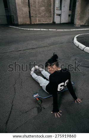 young fit woman with modern haircut sitting on longboard. she is in the shade of building. she wears jeans and sweatshirt. She has tattoos throughout her body. longboard has no prints or aerography