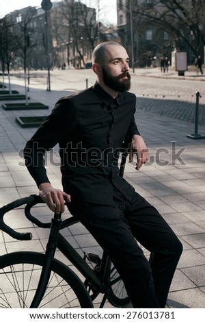 bearded caucasian bald man sits on black fix bicycle against cobbled street . He sits in morning light. There are silhouettes of people. Man is wearing black shirt and trousers and sneakers.