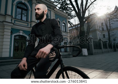bearded caucasian bald man sits on black fix bicycle near old restored building. He sits in morning light in cobbled street. Street is empty. Man is wearing black shirt and trousers and sneakers.