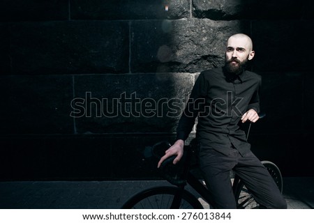 bearded caucasian bald man sits on black fix bicycle against black wall of stone. He sits in thin stripe of light. Street is empty. Man is wearing black shirt and trousers and sneakers.