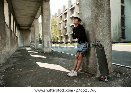 young fit woman with modern haircut standing with longboard. she is in the shade of building. she wears jeans and sweatshirt. She has tattoos throughout her body. longboard has no prints or aerography