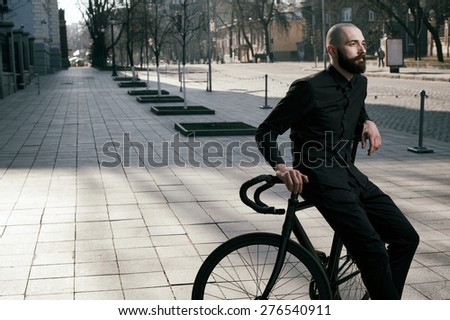 bearded caucasian bald man sits on black fix bicycle against cobbled street . He sits in morning light. There are silhouettes of people. Man is wearing black shirt and trousers and sneakers.