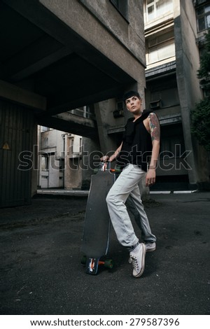 young fit woman with modern haircut standing with longboard. she is in the shade of building. she wears jeans and singlet. She has tattoos throughout her body. longboard has no prints or aerography