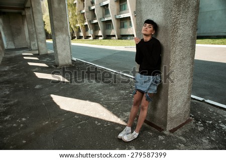young fit woman with modern haircut. she is in the shade of building. she wears jeans and sweatshirt. She has tattoos throughout her body.