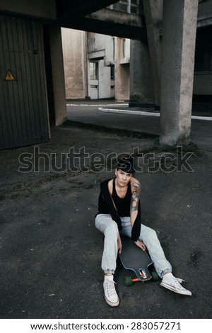 young fit woman with modern haircut sitting on longboard. she is in the shade of building. she wears jeans and singlet. She has tattoos throughout her body. longboard has no prints or aerography