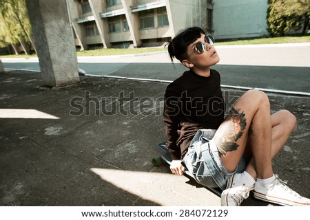 young fit woman with modern haircut sitting on longboard. she is in the shade of building. she wears shorts and sweatshirt. She has tattoos throughout her body. longboard has no prints or aerography