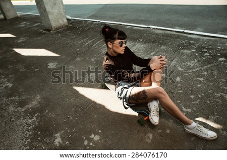 young fit woman with modern haircut sitting on longboard. she is in the shade of building. she wears shorts and sweatshirt. She has tattoos throughout her body. longboard has no prints or aerography