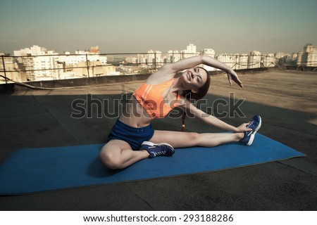 smiling beautiful red haired young lady does stretching on mat on roof of high rise. she wears blue shorts and orange top and blue sneakers. she trains in shade of outbuilding against other high rises