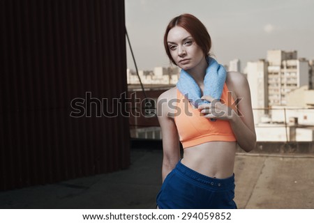 exhausted red haired lady with towel around her neck after workout on roof of high-rise. She wears orange top and blue shorts. Towel is blue. She stands against building and other high rises