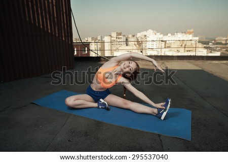 smiling beautiful red haired young woman does stretching on mat on roof of high rise. she wears blue shorts and orange top and blue sneakers. she trains in shade of outbuilding