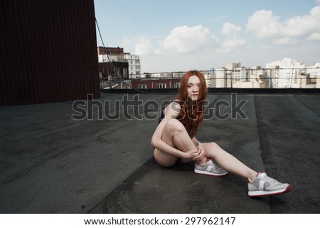 smiling lady with red hair poses on roof of high-rise in purple swimsuit and gray sneakers. she is young and slim. hair fluttering in the wind. she sits against other high-rises and cloudy sky