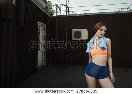 exhausted red haired lady with towel around her neck after workout on roof of high-rise. She wears orange top and blue shorts. She stands against outbuilding and fire escape. She is slim and fit.