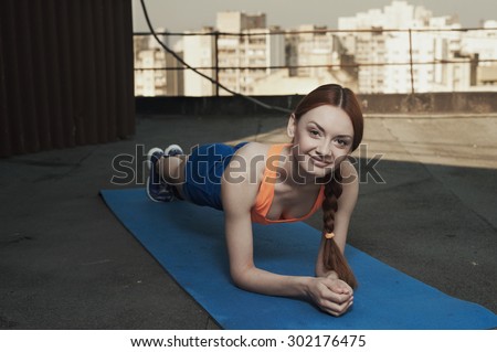 smiling beautiful red haired young woman does the stretching on mat on roof of high rise. she wears blue shorts and orange top and blue sneakers. she trains in shade of outbuilding against high rises