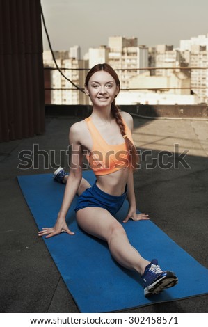 smiling beautiful red haired young woman does the splits on mat on roof of high rise. she wears blue shorts and orange top. she trains in the shade of outbuilding against other high rises