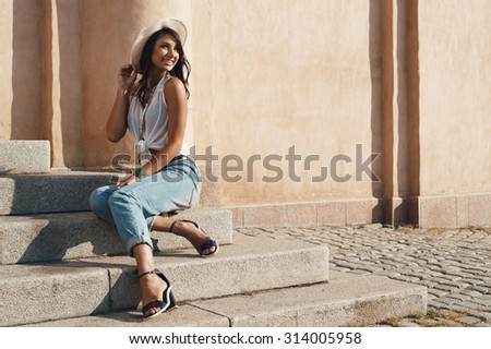 smiling indian lady in jeans, white shirt and white hat against ancient building. She is in harsh morning light. She is positive and playful.