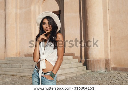 sexy flirting indian lady in jeans, white shirt and white hat against ancient building. She is in harsh morning light. She is positive and playful. Building looks like church or eastern temple