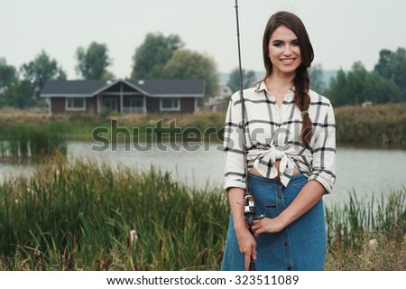 Cute rural brown haired lady posing against ranch house and pond with fishing rod. She stands in grass against rural scape. She wears jeans dress.