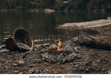 small fireplace with fire near the lake. Two metal bowls near it for gold mining. log on the right for gold digger to warm up near the fire