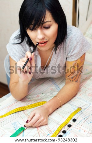 young seamstress with tattoo measuring distance on sewing pattern with tape and pencil