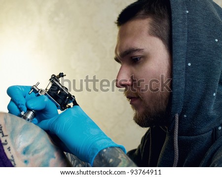 Bearded Caucasian tattooist creates a tattoo on a woman\'s shoulder, close up. He is wearing blue gloves