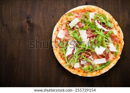 Pizza with prosciutto (parma ham), arugula (salad rocket) and parmesan on dark wooden background top view. Italian cuisine. Space for text.