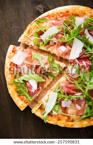 Sliced pizza with prosciutto (parma ham), arugula (salad rocket) and parmesan on dark wooden background top view. Italian cuisine.