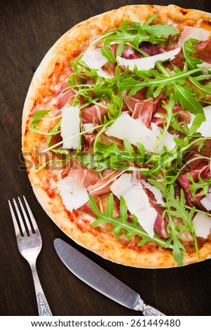 Pizza with prosciutto (parma ham), arugula (salad rocket) and parmesan on dark wooden background top view. Italian cuisine.