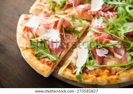 Sliced pizza with prosciutto (parma ham), arugula (salad rocket) and parmesan on dark wooden background close up. Italian cuisine.