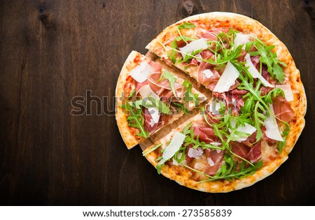 Sliced pizza with prosciutto (parma ham), arugula (salad rocket) and parmesan on dark wooden background top view. Italian cuisine. Space for text.