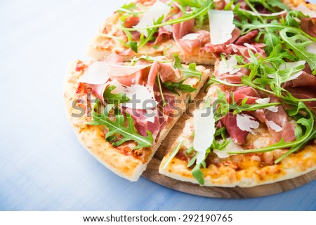 Sliced pizza with prosciutto (parma ham), arugula (salad rocket) and parmesan on blue wooden background close up. Italian cuisine.