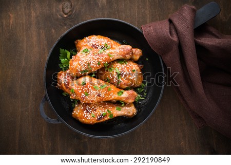 Baked spicy chicken legs with sesame and parsley in cast iron frying pan on dark wooden background top view.