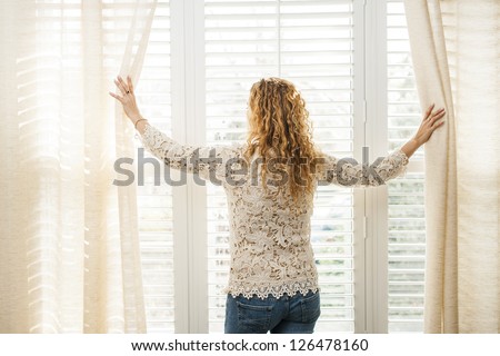 Woman looking out big bright window with curtains and blinds