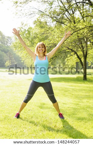 Female fitness instructor doing jumping jacks exercising in green park
