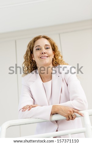 Smiling proud business woman standing in office hallway leaning on railing shot from below