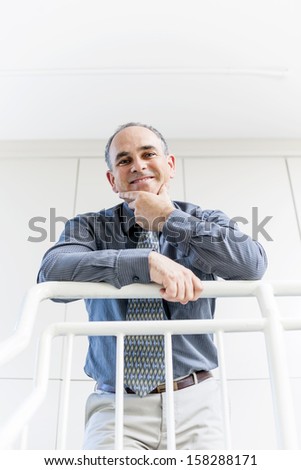 Smiling proud business man standing in office hallway leaning on railing shot from below