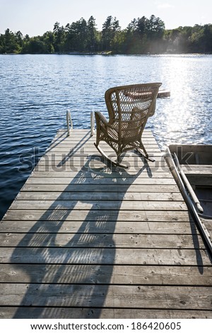 Wicker rocking chair on wooden dock in summer at small lake casting long shadow