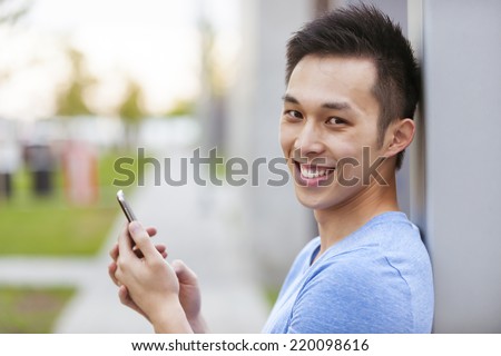 Smiling young asian man holding smart phone and looking at camera on city sidewalk