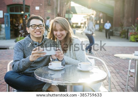 Two laughing young people looking into smartphone while sitting at a table in outdoor cafe