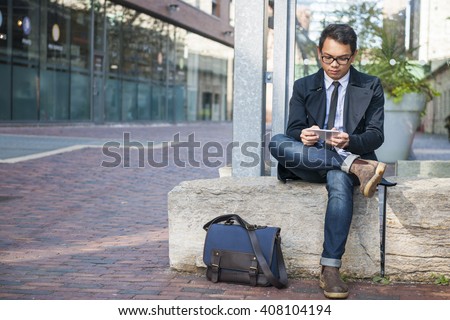 Young serious asian business man looking at mobile phone sitting outside on city street with copy space