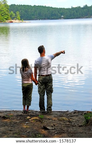 Father and daughter standing on the lake shore and looking at calm water, father pointing out something in the distance