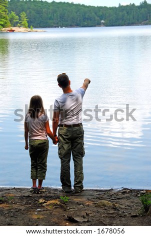 Father and daughter standing on the lake shore and looking at calm water, father pointing at something on the other shore
