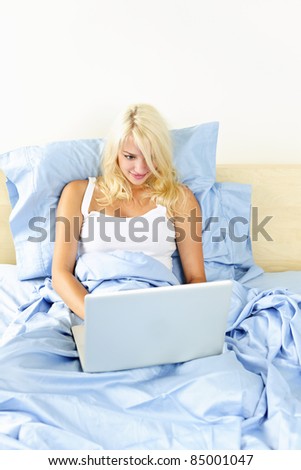 Happy blonde woman using computer sitting in bed