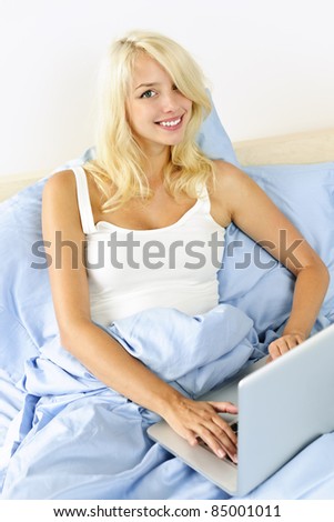 Happy blonde woman using laptop computer at home sitting in bed