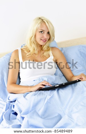 Happy blonde woman using tablet computer sitting in bed at home