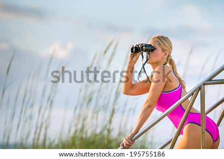 lovely female in pink one-piece swimsuit scans the ocean with binoculars at a florida beach