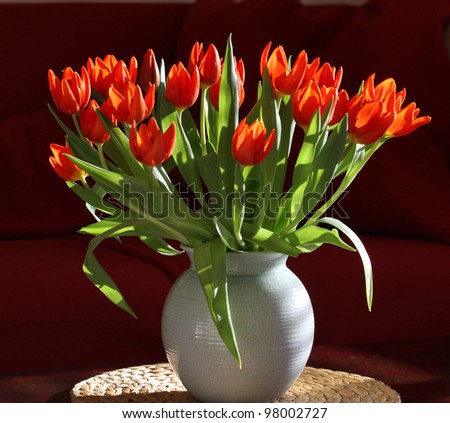 Vase with Orange Tulips in Modern Interior. Crackle-glazed vase, round place-mat of water hyacinth; background of burgundy-red sofa.